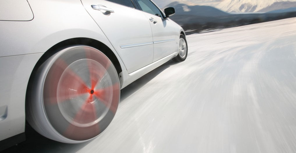AutoSock mounted on rear wheels of a white car, driving on snow towards a snowy mountain