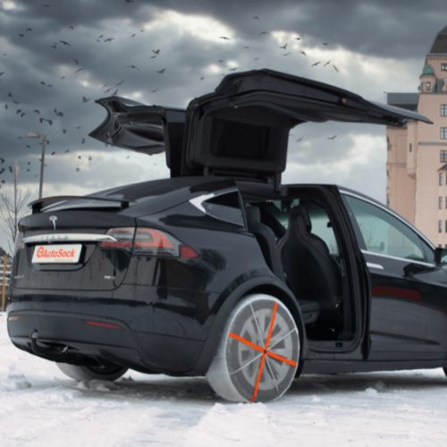 AutoSock tire socks mounted on rear wheels of a Tesla Y passenger car, standing on snow with open wing doors